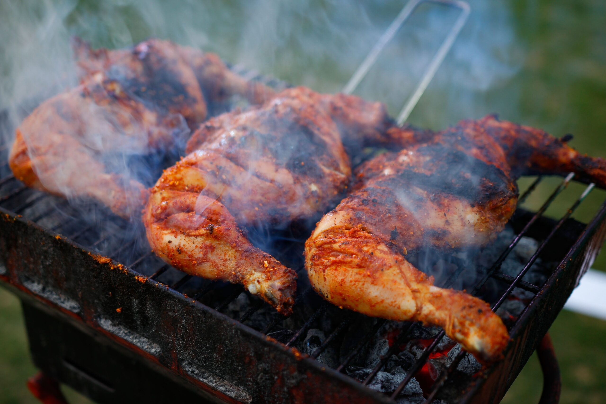 Close-up of spicy chicken grilling on a charcoal barbecue with visible smoke outdoors.